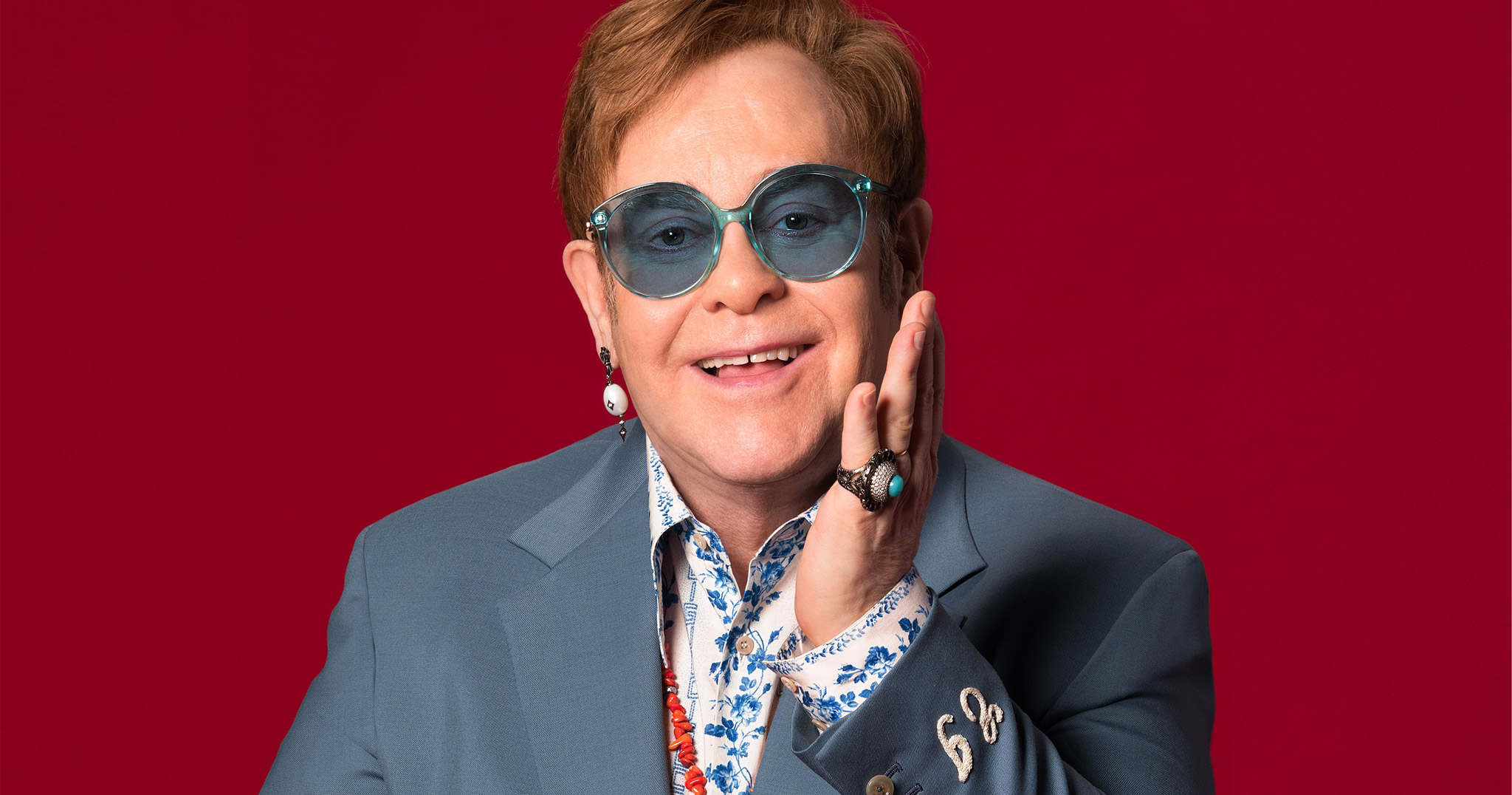 Elton John in blue glasses against a red background.
